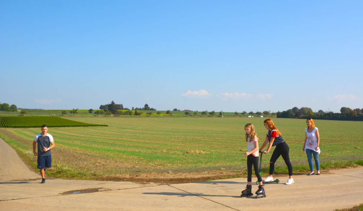 Menschen auf einem Feldweg im Schmidener Feld, eine Person läuft, zwei fahren Roller und Inlineskates. Im Hintergrund weite Felder und blauer Himmel., © Remstal Tourismus e.V.