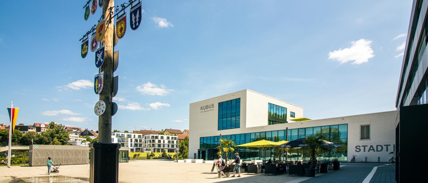 Moderner Marktplatz in Remseck mit dem Gebäude 'KUBUS', einem Maibaum mit Wappen und Menschen, die spazieren gehen. Sonniger Tag mit blauem Himmel., © Stuttgart-Marketing GmbH, Sarah Schmid Moderner Marktplatz in Remseck mit dem Gebäude 'KUBUS', einem Maibaum mit Wappen und Menschen, die spazieren gehen. Sonniger Tag mit blauem Himmel., © Stuttgart-Marketing GmbH, Sarah Schmid