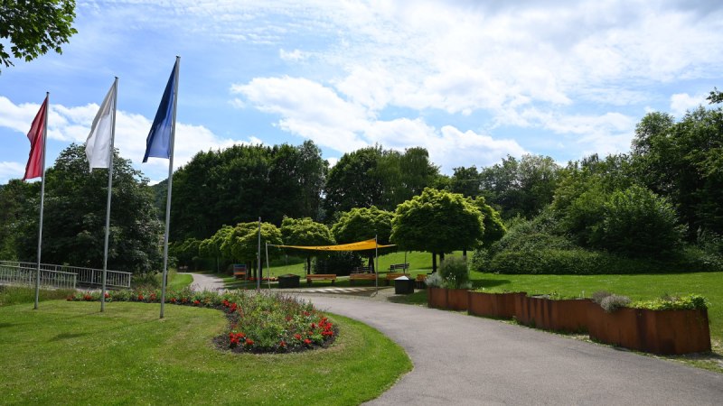Der Kurpark Bad Urach zeigt einen Weg mit Blumenbeeten, drei Fahnen und B&auml;umen unter einem blauen Himmel mit Wolken.