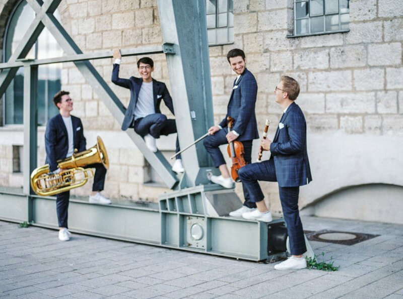 Vier Männer in Anzügen mit Instrumenten posieren lächelnd vor einer Steinmauer. Sie wirken entspannt und fröhlich., © Kammerkonzerte Schwäbisch Hall e.V.