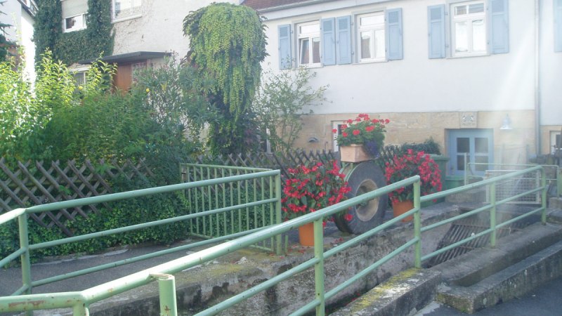 Haus mit blauen Fensterläden, umgeben von Pflanzen und Blumen. Ein grünes Geländer führt über einen kleinen Graben., © Natur.Nah. Schönbuch & Heckengäu