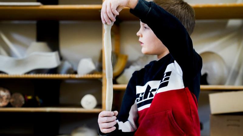 Ein Junge in einem rot-schwarzen Pullover formt Ton in einem Workshop. Im Hintergrund sind Regale mit Materialien zu sehen., © Kunstschule Labyrinth Ein Junge in einem rot-schwarzen Pullover formt Ton in einem Workshop. Im Hintergrund sind Regale mit Materialien zu sehen., © Kunstschule Labyrinth