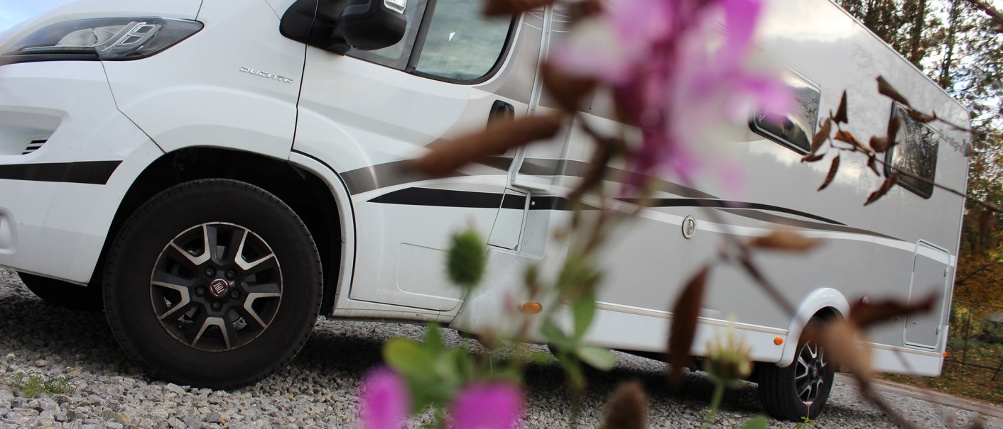 Weißes Wohnmobil auf Kiesplatz, im Vordergrund unscharfe Blumen. Seitliche Ansicht mit Fokus auf die Räder., © Erlebnisregion Schwäbischer Albtrauf e. V.