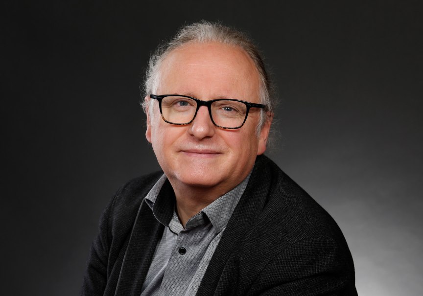 Ein Mann mit Brille und grauem Haar l&auml;chelt in die Kamera. Er tr&auml;gt ein dunkles Sakko und ein Hemd. Der Hintergrund ist dunkel und schlicht.