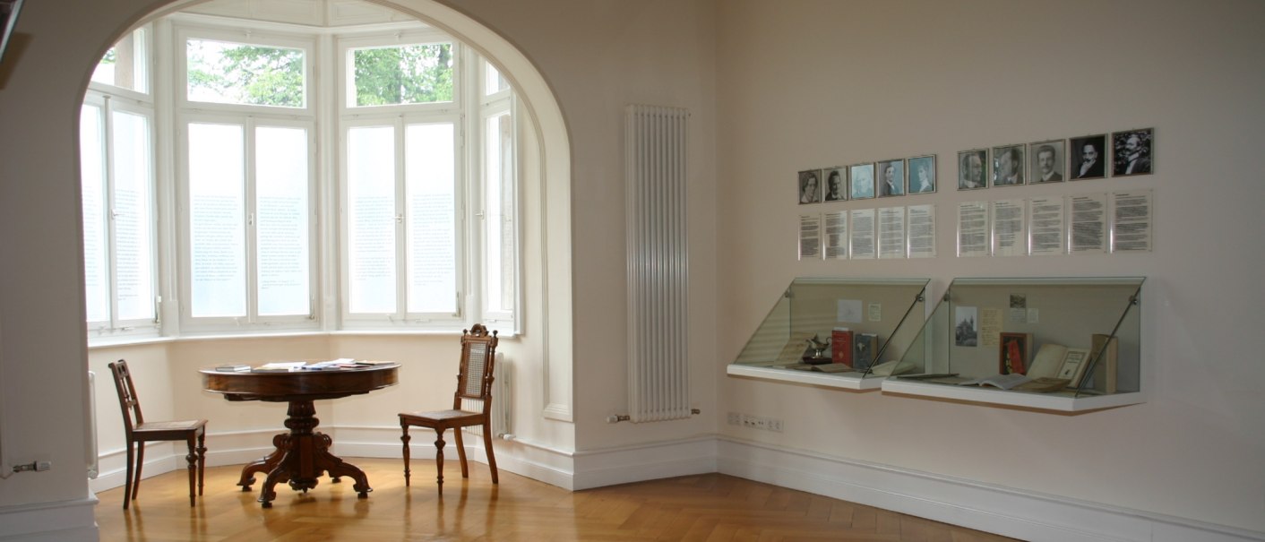 Ein heller Raum mit Erkerfenster, rundem Tisch und St&uuml;hlen. An der Wand h&auml;ngen Fotos und Texte, daneben Vitrinen mit B&uuml;chern., &copy; Gemeindearchiv Bad Boll