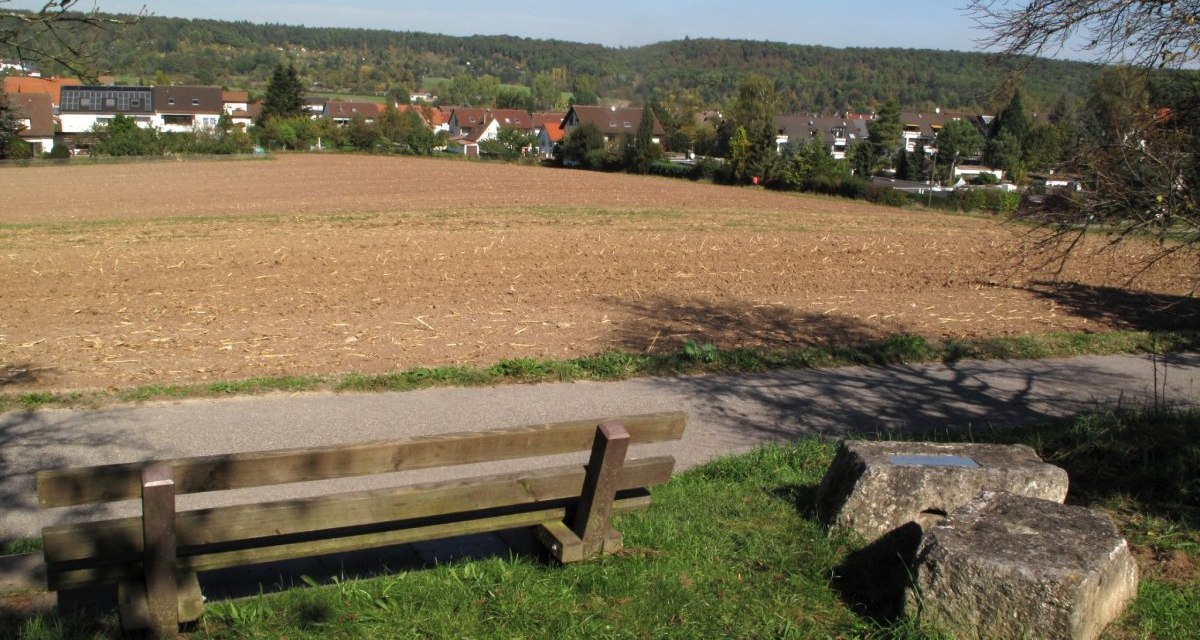 Eine Holzbank steht vor einem Feld, dahinter sind Häuser und ein bewaldeter Hügel zu sehen. Der Himmel ist klar und blau., © RadL Leonberg Eine Holzbank steht vor einem Feld, dahinter sind Häuser und ein bewaldeter Hügel zu sehen. Der Himmel ist klar und blau., © RadL Leonberg