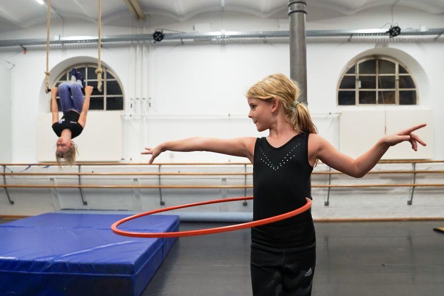 Zwei Kinder üben Zirkuskunststücke: Eines hängt kopfüber an einem Seil, das andere spielt mit einem Hula-Hoop-Reifen. Hintergrund: Turnraum., © Kunstschule Labyrinth Zwei Kinder üben Zirkuskunststücke: Eines hängt kopfüber an einem Seil, das andere spielt mit einem Hula-Hoop-Reifen. Hintergrund: Turnraum., © Kunstschule Labyrinth