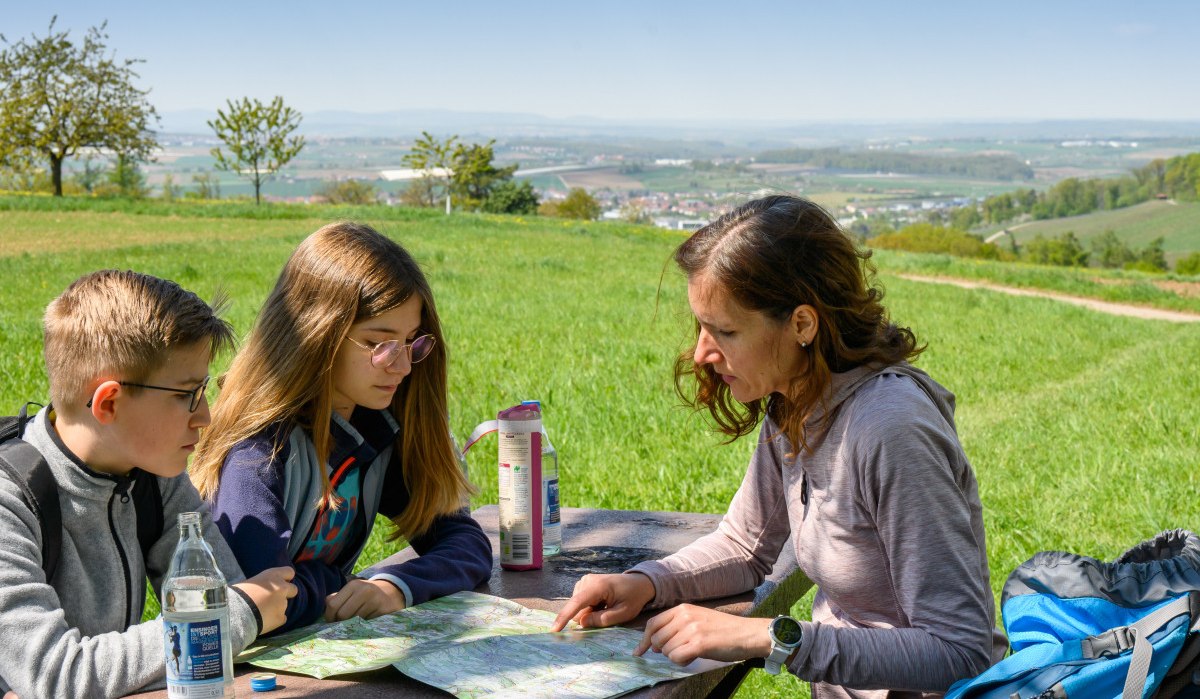 Drei Personen sitzen an einem Tisch im Freien und betrachten eine Karte. Im Hintergrund ist eine weite Landschaft mit Feldern und Bäumen zu sehen., © Stadtverwaltung Winnenden