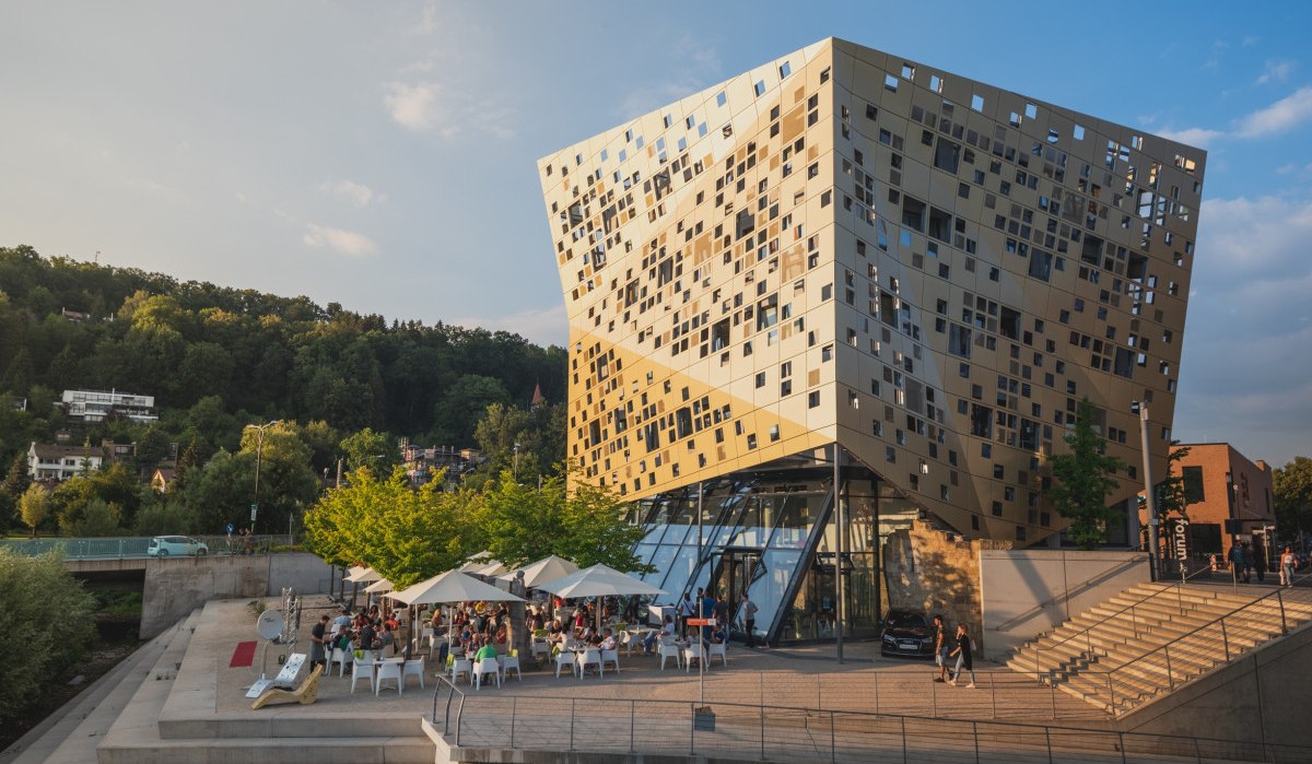 Modernes Gebäude mit goldener Fassade, viele Fenster. Menschen sitzen unter Sonnenschirmen. Umgeben von Bäumen und Treppen., © Remstal Tourismus e.V. I Architekt: isin Architekten