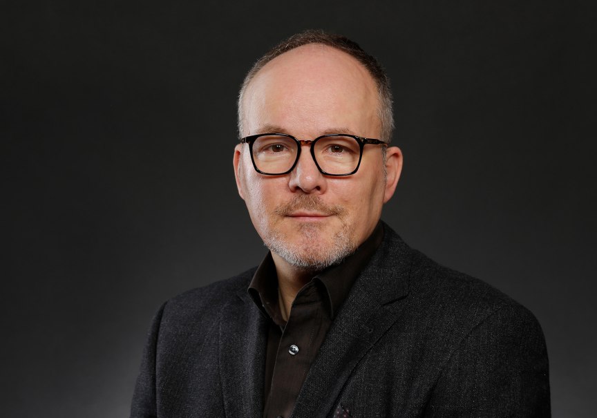 Ein Mann mit Brille und Bart in einem dunklen Anzug vor einem neutralen, dunklen Hintergrund.