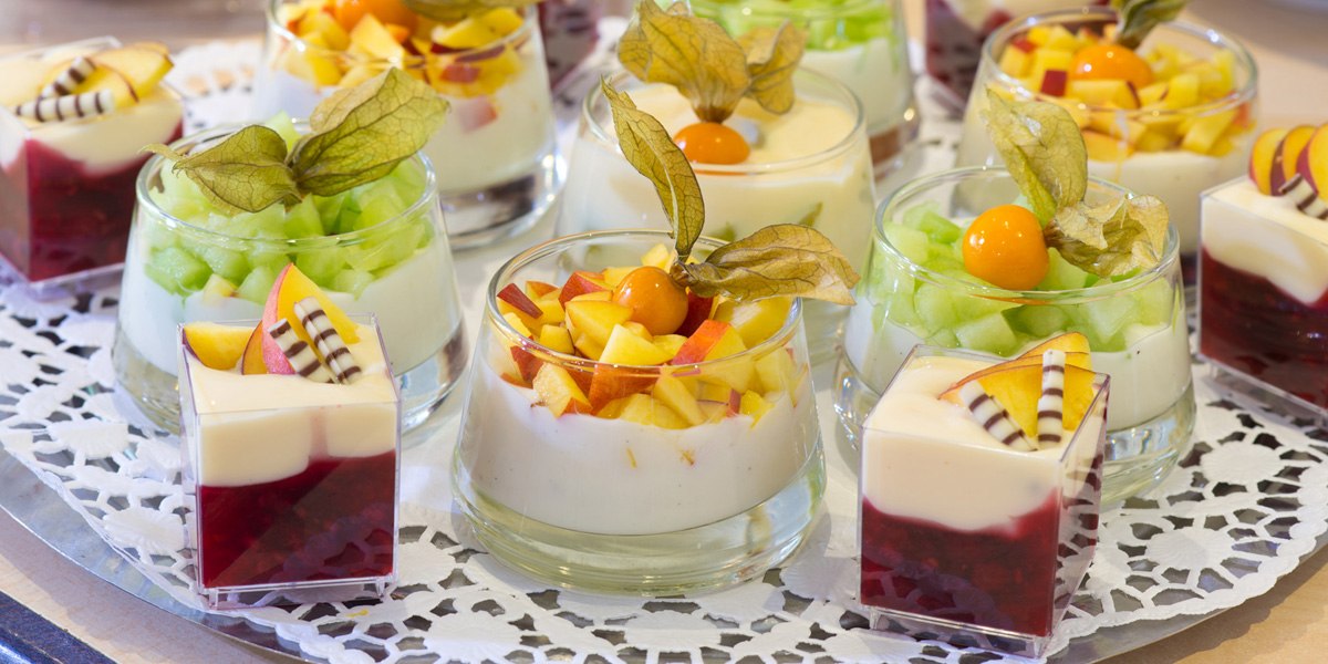 Verschiedene Dessertgläser mit Joghurt, frischem Obst und Physalis auf einem dekorativen Tablett arrangiert., © ACHAT Hotels Deutschland