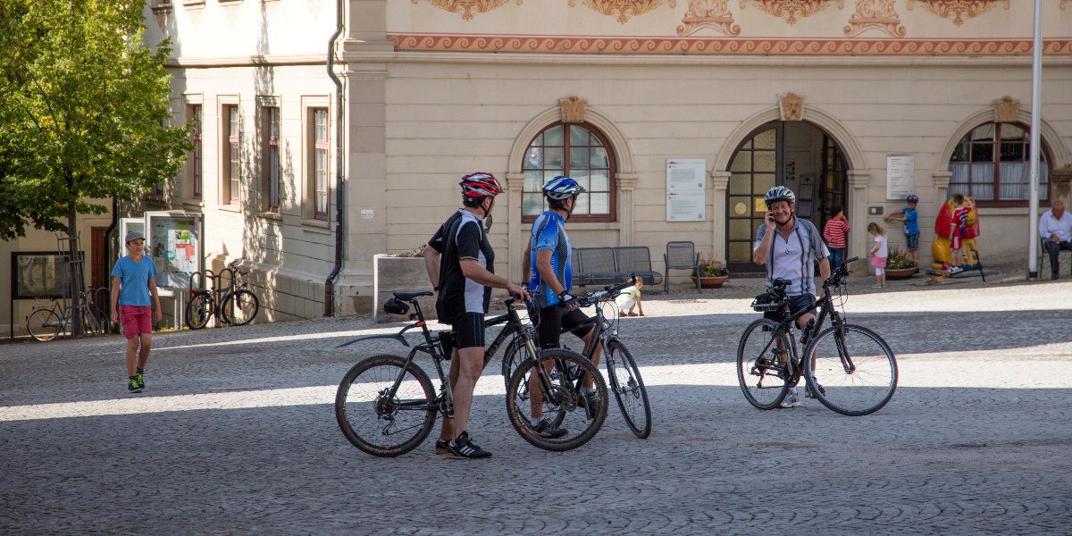 Drei Radfahrer unterhalten sich auf einem gepflasterten Platz vor einem historischen Gebäude. Ein Kind läuft vorbei, im Hintergrund sind weitere Personen zu sehen., © Stuttgart-Marketing GmbH Drei Radfahrer unterhalten sich auf einem gepflasterten Platz vor einem historischen Gebäude. Ein Kind läuft vorbei, im Hintergrund sind weitere Personen zu sehen., © Stuttgart-Marketing GmbH