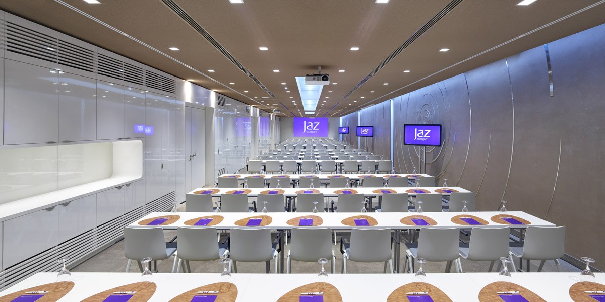 Moderner Konferenzraum mit weißen Tischen und Stühlen, Bildschirmen an den Wänden und einem Projektor an der Decke., © Jaz Hotel Stuttgart