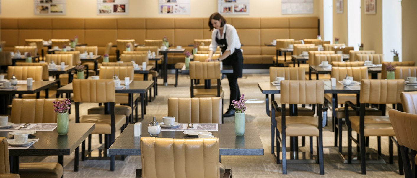 Ein Restaurant mit beige gepolsterten St&uuml;hlen und gedeckten Tischen. Eine Person richtet einen Tisch her., &copy; Arcotel Hotels
