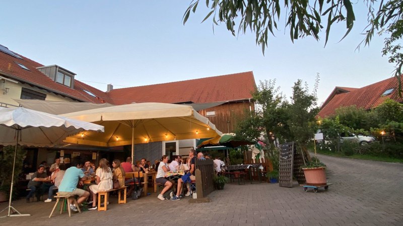 Menschen sitzen unter gro&szlig;en Sonnenschirmen in einem gem&uuml;tlichen Hof mit roten D&auml;chern und B&auml;umen., &copy; Weinhof Zai&szlig;