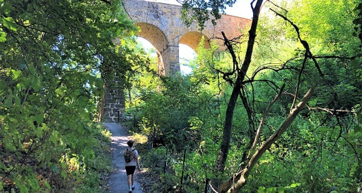 Ein Wanderer auf einem schattigen Pfad unter einer steinernen Bogenbrücke, umgeben von dichtem Grün., © Touristik und Marketing GmbH Ein Wanderer auf einem schattigen Pfad unter einer steinernen Bogenbrücke, umgeben von dichtem Grün., © Touristik und Marketing GmbH