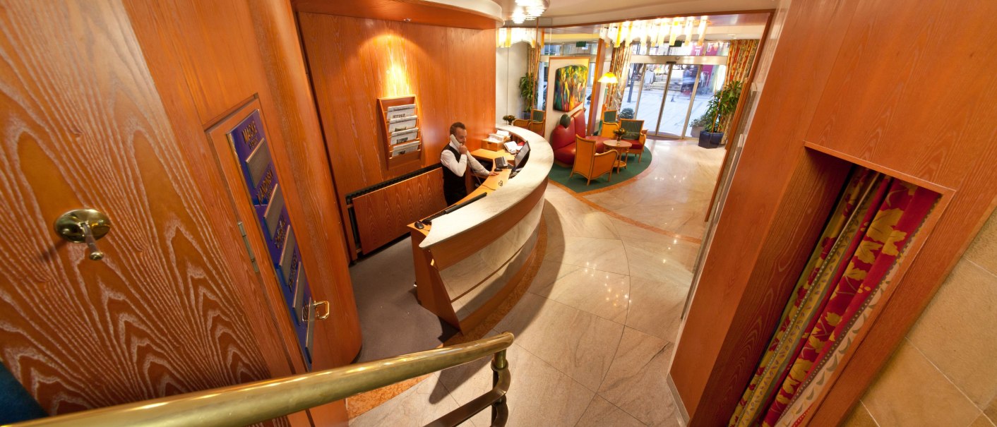 Hotelrezeption mit einem Empfangstresen, an dem ein Mitarbeiter telefoniert. Im Hintergrund ein Sitzbereich mit Sesseln und Glastüren., © Novum Boulevard Stuttgart City