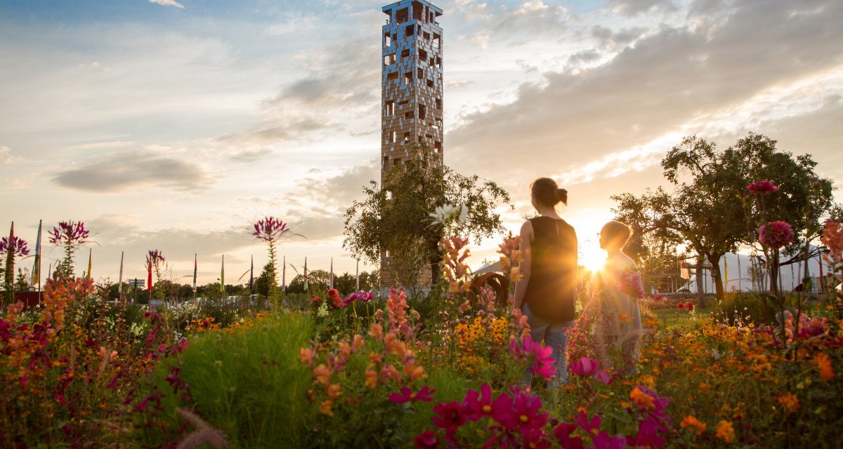 Ein Turm im Himmelsgarten, umgeben von bunten Blumen. Zwei Personen stehen im Vordergrund, während die Sonne im Hintergrund untergeht., © Foto Thomas Zehnder