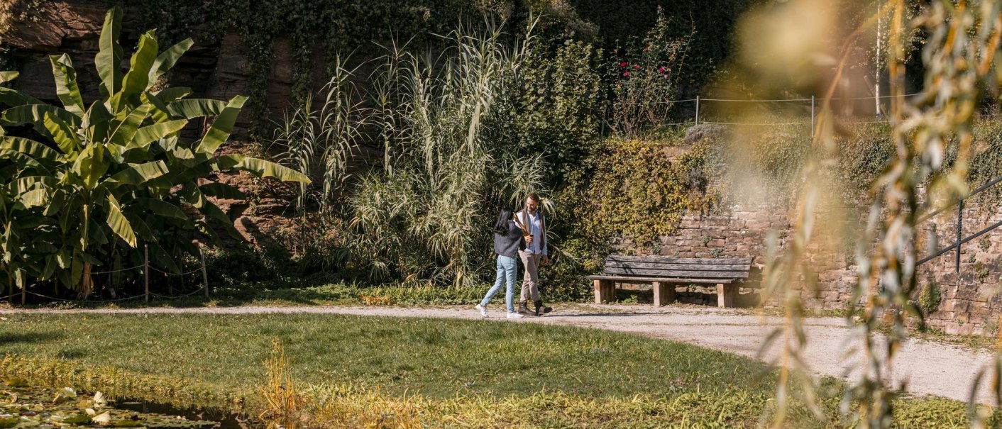 Zwei Personen spazieren in einem Garten mit Bananenpflanzen, Steinmauer und Bank. Im Vordergrund sind Gräser und ein Teich zu sehen., © Stuttgart-Marketing GmbH, Sarah Schmid