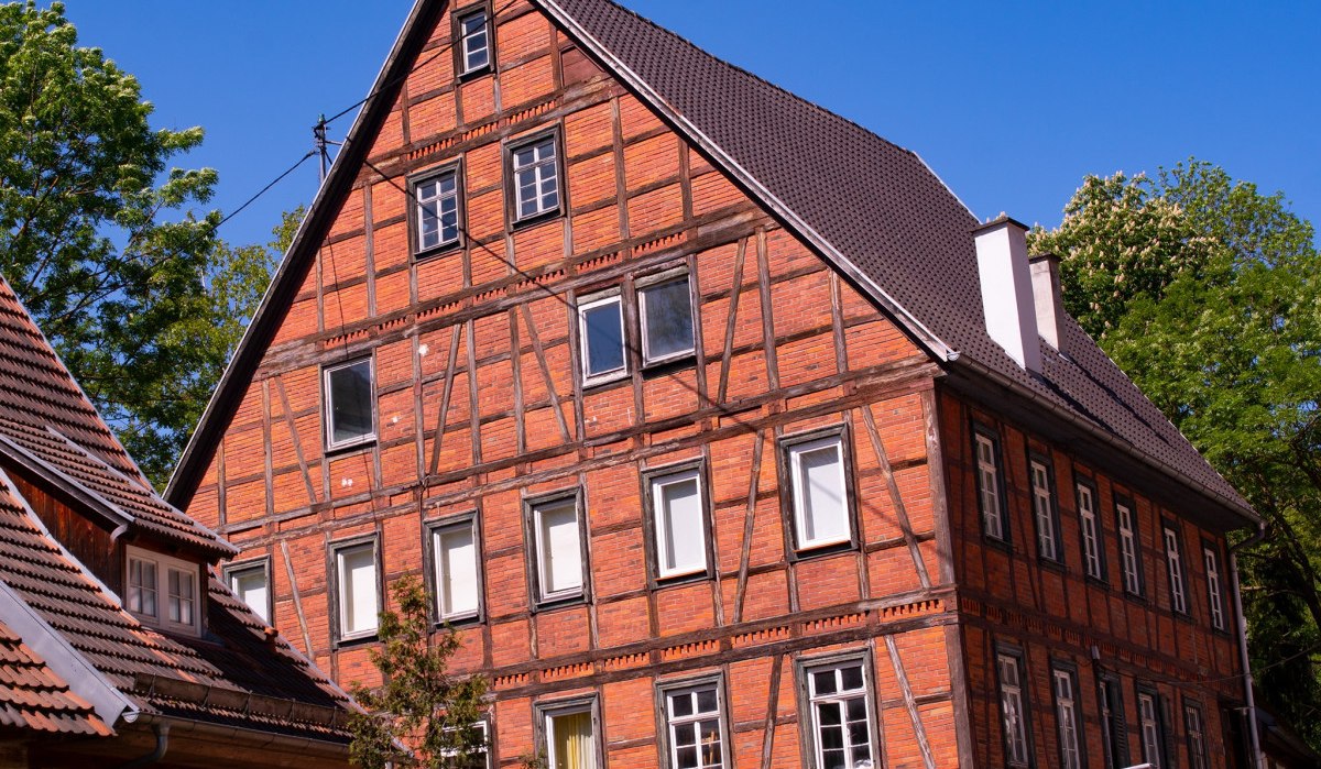 Ein großes Fachwerkhaus mit rotem Ziegelmauerwerk und dunklem Dach steht vor einem klaren blauen Himmel. Bäume umgeben das Gebäude., © Agentur Arcos Ein großes Fachwerkhaus mit rotem Ziegelmauerwerk und dunklem Dach steht vor einem klaren blauen Himmel. Bäume umgeben das Gebäude., © Agentur Arcos