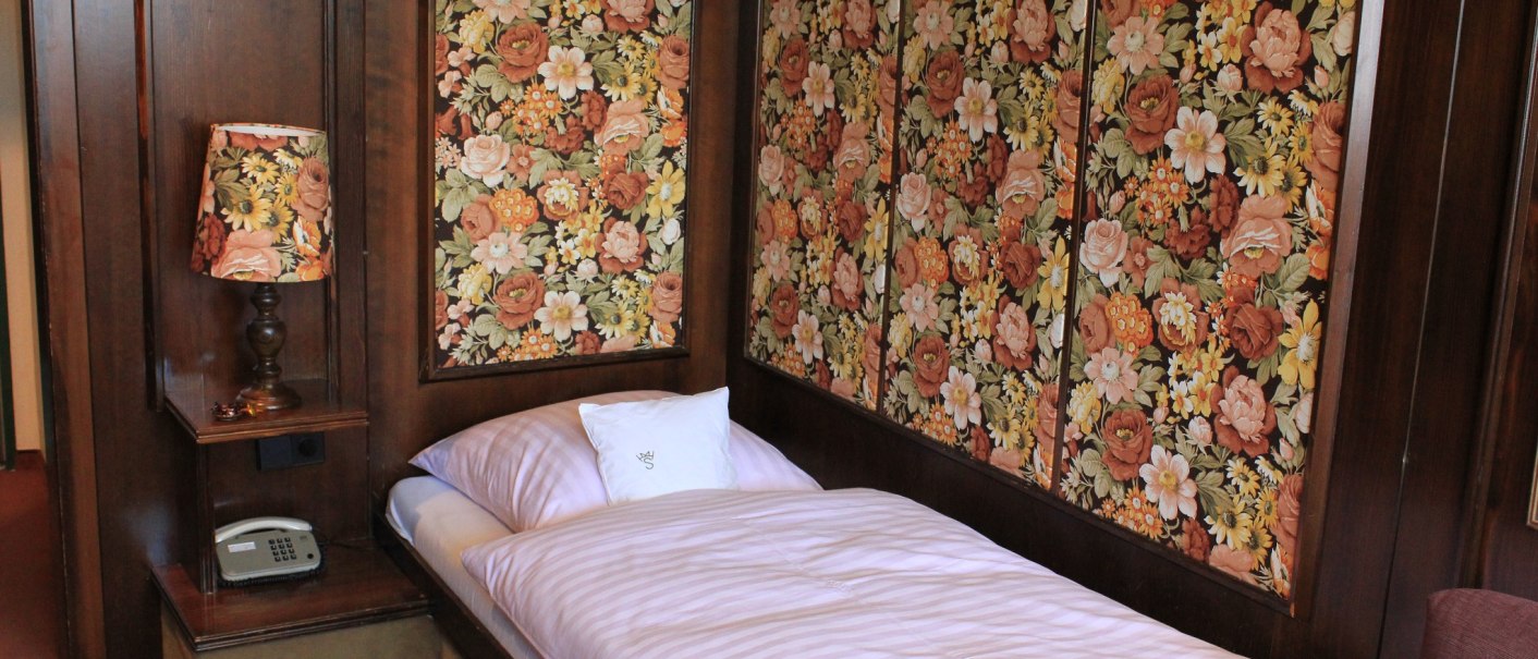 Ein Hotelzimmer mit einem Einzelbett, floraler Tapete und passender Lampe. Ein Telefon steht auf einem kleinen Regal., © Hotel Spahr Ein Hotelzimmer mit einem Einzelbett, floraler Tapete und passender Lampe. Ein Telefon steht auf einem kleinen Regal., © Hotel Spahr