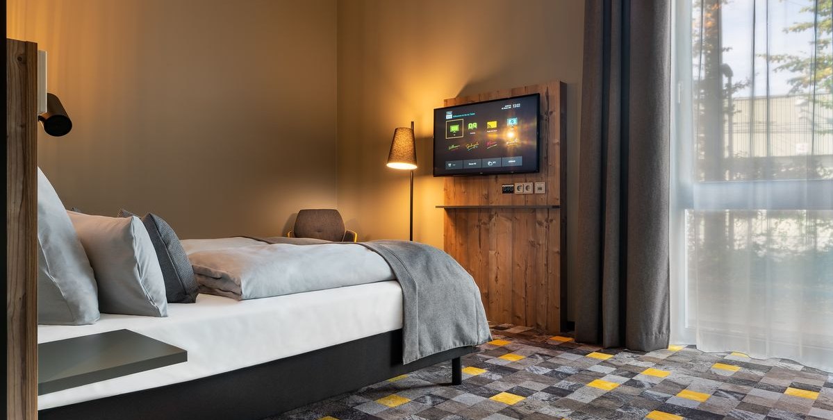 Modernes Hotelzimmer mit einem Bett, einem an der Wand montierten Fernseher und einer Stehlampe. Der Boden hat ein geometrisches Muster., © Novum Hospitality Modernes Hotelzimmer mit einem Bett, einem an der Wand montierten Fernseher und einer Stehlampe. Der Boden hat ein geometrisches Muster., © Novum Hospitality