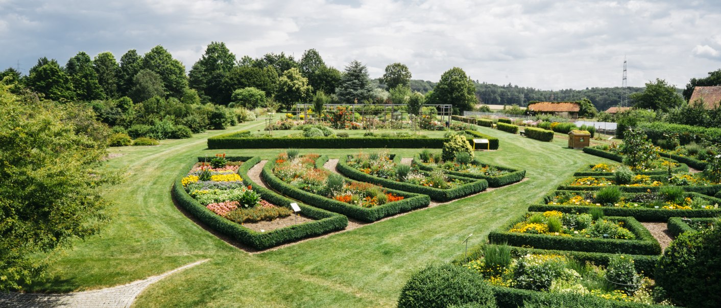 Ein weitläufiger Garten mit symmetrischen Blumenbeeten und gepflegten Hecken, umgeben von Bäumen und einem blauen Himmel., © HfWU, Manuel stark