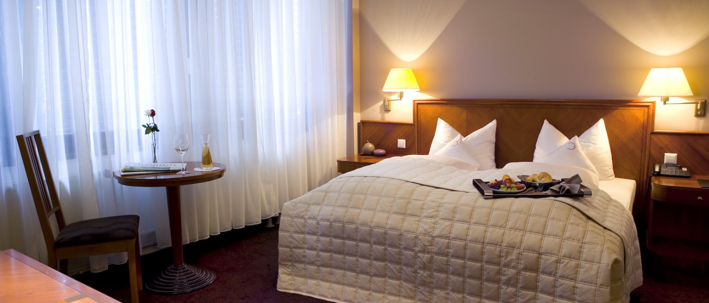 Gemütliches Hotelzimmer mit Doppelbett, Nachttischlampen, kleinem Tisch und Stuhl am Fenster. Ein Tablett mit Frühstück steht auf dem Bett., © Ross Messehotel Gemütliches Hotelzimmer mit Doppelbett, Nachttischlampen, kleinem Tisch und Stuhl am Fenster. Ein Tablett mit Frühstück steht auf dem Bett., © Ross Messehotel
