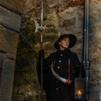 Ein Nachtwächter in historischer Kleidung mit Laterne und Hellebarde steht vor einer alten Steinmauer., © Stadt Besigheim
