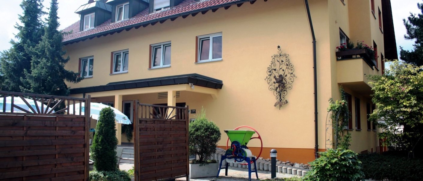 Gelbes Gebäude mit rotem Dach und Hotelaufschrift. Vor dem Eingang ein Holzzaun und Pflanzen. Dekoratives Wandornament und grüne Maschine im Vordergrund., © TOMAS Gelbes Gebäude mit rotem Dach und Hotelaufschrift. Vor dem Eingang ein Holzzaun und Pflanzen. Dekoratives Wandornament und grüne Maschine im Vordergrund., © TOMAS