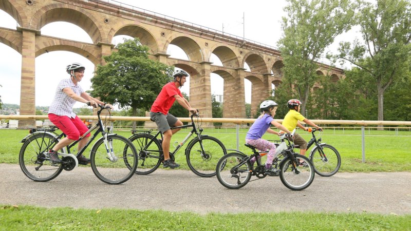 Eine Familie fährt mit Fahrrädern auf einem Weg vor dem Bietigheimer Viadukt. Im Hintergrund sind Bäume und das Viadukt zu sehen., © Land der 1000 Hügel - Kraichgau-Stromberg
