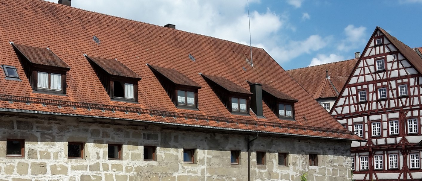 langestrecktes Steingebäude mit kleinen Fenstern und rotbraunem Ziegeldach mit mehreren Dachgauben, im Hintergrund rechts der Fachwerkgiebel des Alten Schlosses mit braunem Holz und weißen Gefachen, © Stadt Gaildorf