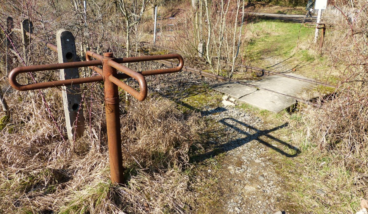 Ein rostiges Drehkreuz steht neben einem Pfad, der &uuml;ber die Schienen einer stillgelegten Bahnstrecke f&uuml;hrt. Die Umgebung ist von Gras und B&uuml;schen bewachsen., &copy; Natur.Nah. Sch&ouml;nbuch & Heckeng&auml;u