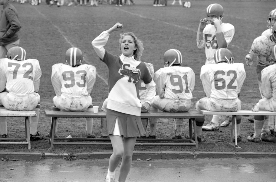 Ein Cheerleader in Uniform feuert ein Football-Team an, das auf einer Bank am Spielfeldrand sitzt. Die Spieler tragen Helme und schmutzige Trikots., &copy; Ludwigsburg Museum
