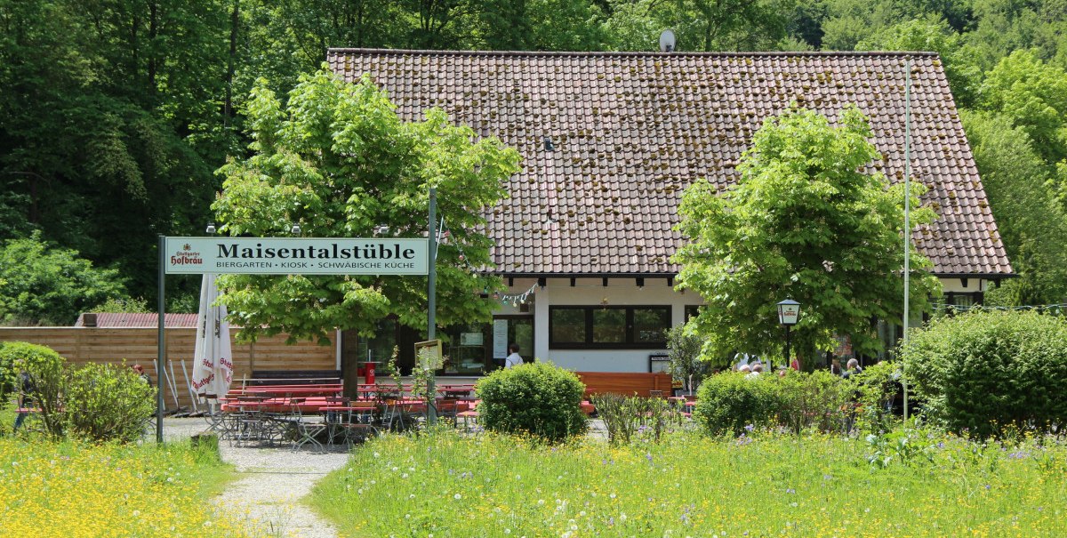 Biergarten Maisentalstüble mit roten Tischen und Stühlen, umgeben von grüner Natur und blühenden Wiesen.
