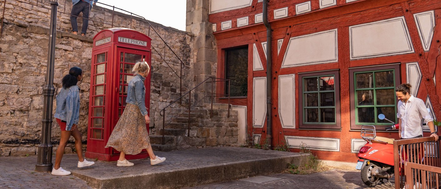 Zwei Personen gehen an einer roten Telefonzelle vorbei, neben einem Fachwerkhaus. Ein Mann steht bei einem Roller. Eine Person geht die Treppe hinauf., © SMG, Martina Denker