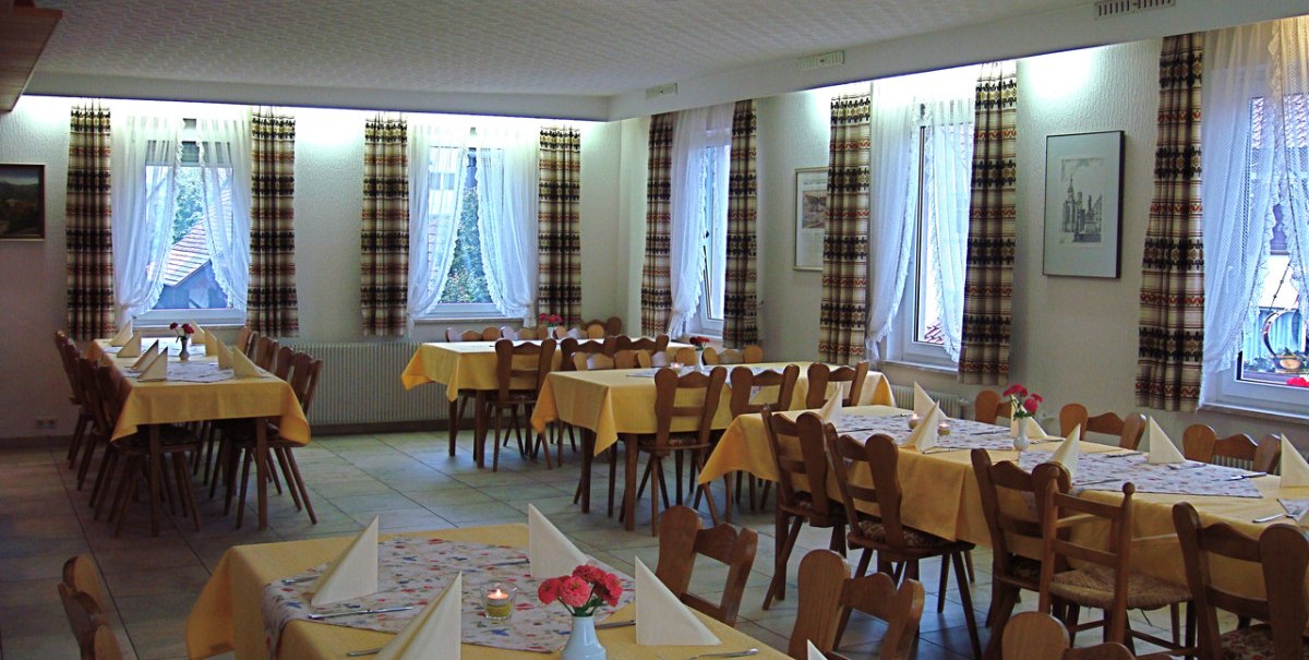 Ein Speisesaal mit gelben Tischdecken, Holzstühlen und gefalteten Servietten. Große Fenster mit karierten Vorhängen lassen Tageslicht herein.