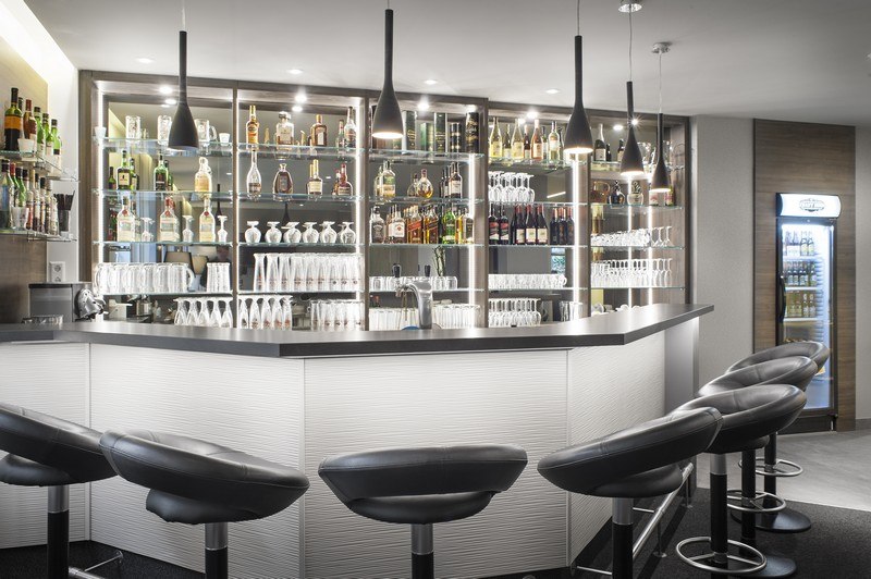 Moderne Bar mit schwarzen Hockern, beleuchteten Regalen voller Flaschen und Gläser, und stilvollen Hängelampen., © TOMAS