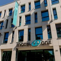 Moderne Fassade eines Gebäudes mit Motel One Logo und blauer Fahne, die im Wind weht. Klare Linien und große Fenster prägen das Design., © Motel One Stuttgart-Bad Cannstatt