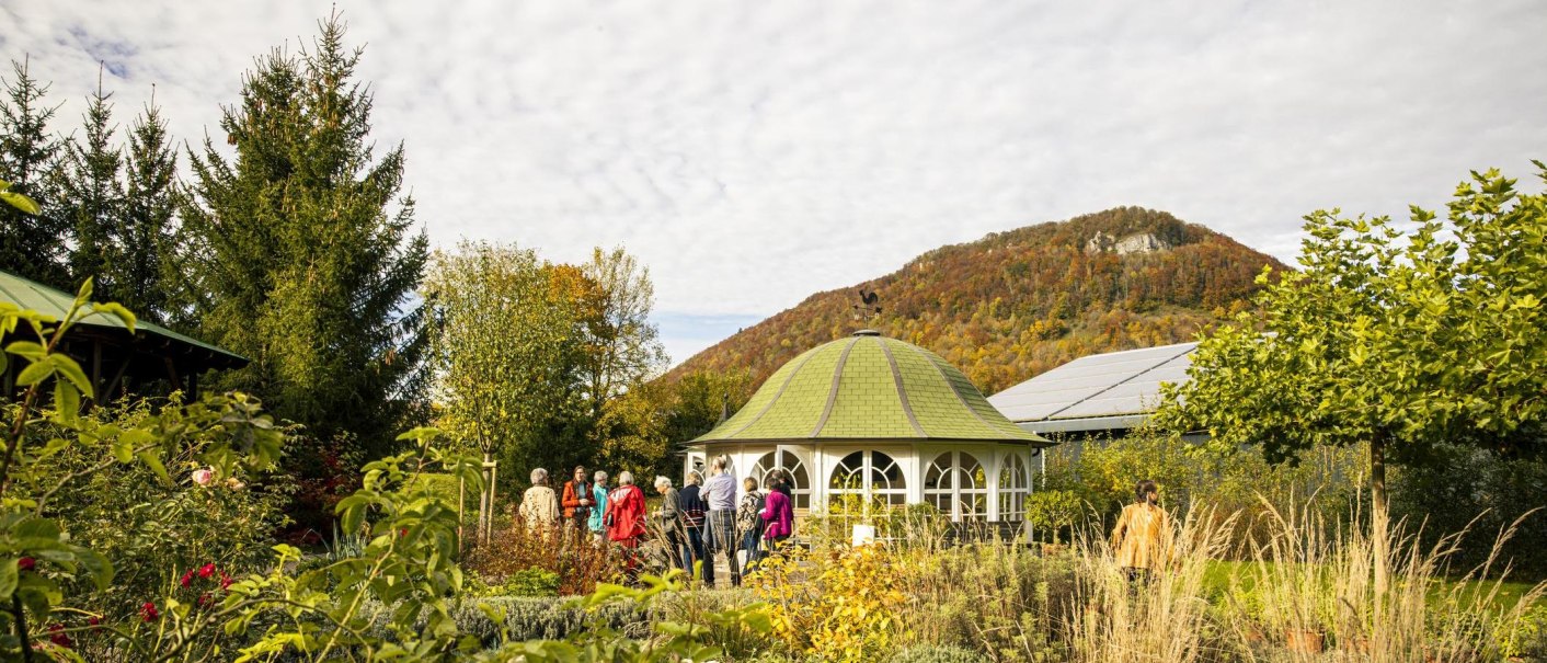 Menschen stehen in einem Kräutergarten vor einem Pavillon. Im Hintergrund ist ein bewaldeter Hügel zu sehen. Der Himmel ist bewölkt., © SMG, Sarah Schmid