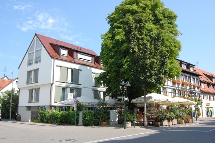 Moderne und traditionelle Gebäude an einer Straßenecke, mit einem großen Baum und einem Café mit Sonnenschirmen., © Jeanette Bender Moderne und traditionelle Gebäude an einer Straßenecke, mit einem großen Baum und einem Café mit Sonnenschirmen., © Jeanette Bender