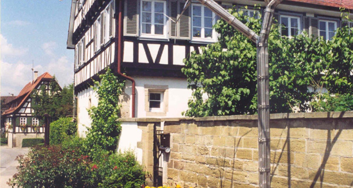 Ein Brunnen mit einer Metallskulptur in Form eines Baumes steht vor einem traditionellen Fachwerkhaus. Bunte Blumen und eine Bank sind im Vordergrund., &copy; Alfred R&ouml;sner