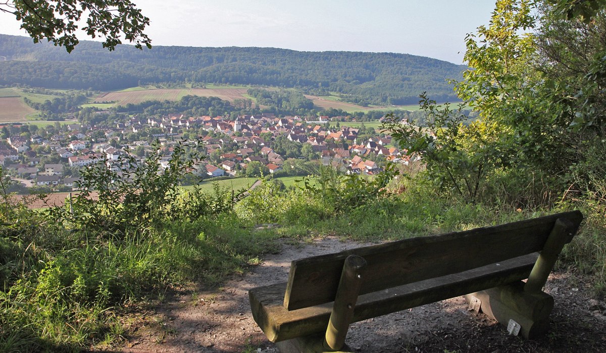 Eine Holzbank bietet einen malerischen Blick auf das Dorf Gündelbach, umgeben von grünen Feldern und bewaldeten Hügeln., © Land der 1000 Hügel - Kraichgau-Stromberg Eine Holzbank bietet einen malerischen Blick auf das Dorf Gündelbach, umgeben von grünen Feldern und bewaldeten Hügeln., © Land der 1000 Hügel - Kraichgau-Stromberg