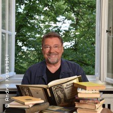 Ein Mann sitzt l&auml;chelnd vor einem offenen Fenster, umgeben von B&uuml;chern. Drau&szlig;en sind gr&uuml;ne B&auml;ume zu sehen. &copy; Andr&eacute; Kowalski, &copy; links im Bild