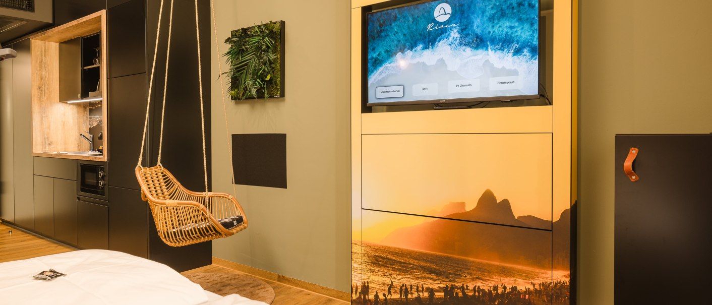 Modernes Hotelzimmer mit Hängesessel, Fernseher mit Strandbild, kleiner Küchenzeile und dekorativem Pflanzenbild an der Wand., © TOMAS Modernes Hotelzimmer mit Hängesessel, Fernseher mit Strandbild, kleiner Küchenzeile und dekorativem Pflanzenbild an der Wand., © TOMAS