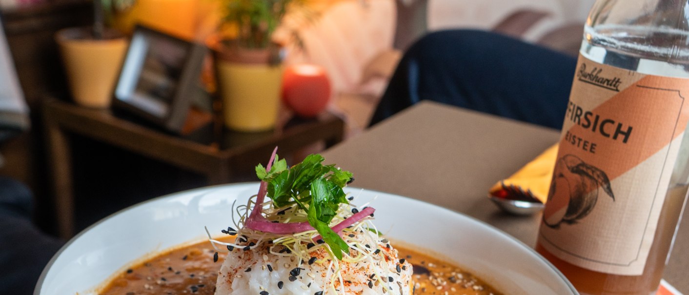 Ein Teller mit Reis und Curry, garniert mit Sesam und Kr&auml;utern, steht auf einem Tisch. Im Hintergrund sind Pflanzen und ein Wandbild mit Tukanen zu sehen., &copy; HerzoginN&uuml;rtingen