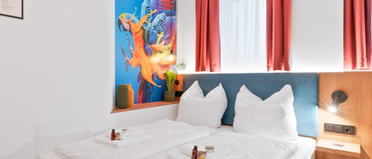 Modernes Doppelzimmer mit weißem Bett, Papageien-Wandbild, roten Vorhängen und gelbem Hocker. Pflegeprodukte auf dem Bett., © JUNGLE N°5 GmbH