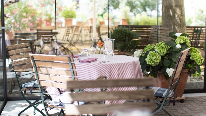 Ein einladend gedeckter Tisch mit rot-wei&szlig; gestreifter Tischdecke in einem Gartenrestaurant. Umgeben von Holzm&ouml;beln und bl&uuml;henden Pflanzen., &copy; Valentino