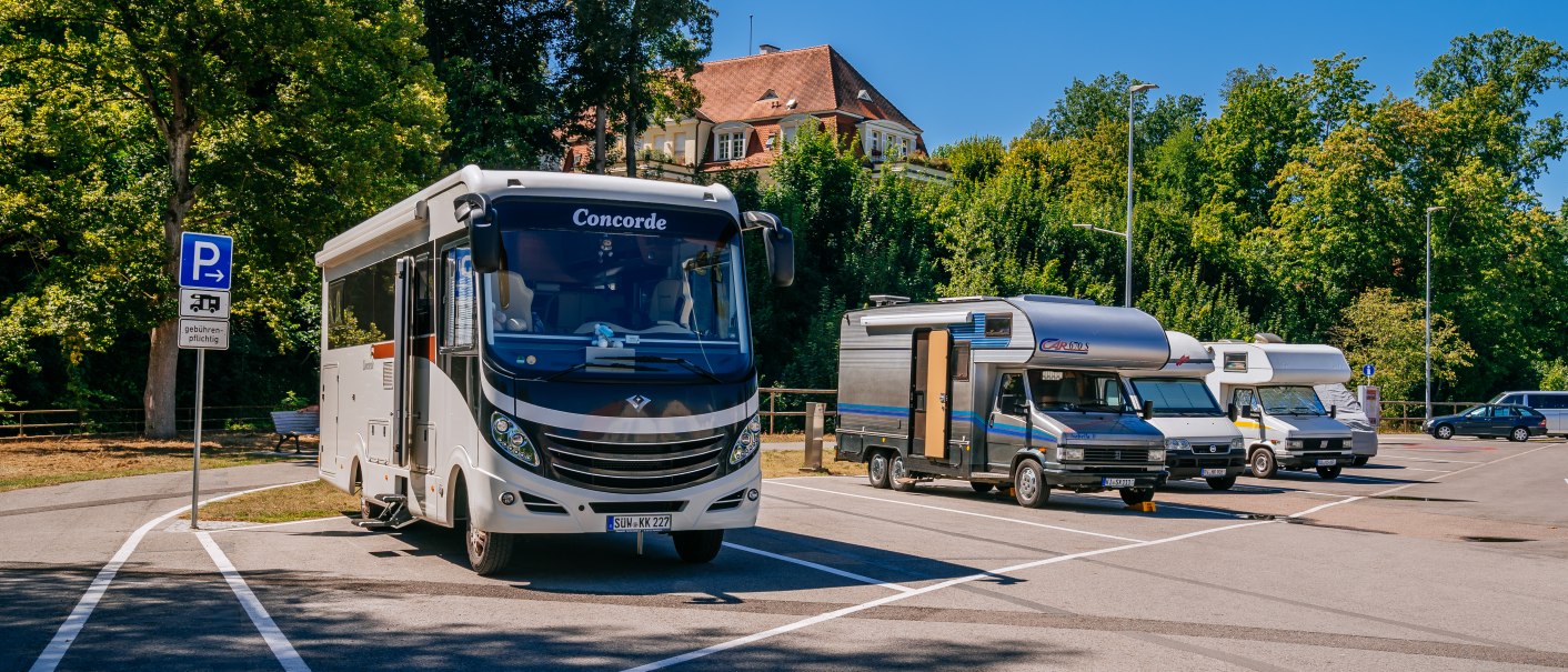 Wohnmobilstellplatz mit mehreren Wohnmobilen unter blauem Himmel, umgeben von Bäumen. Ein Schild weist auf gebührenpflichtiges Parken hin., © SMG, Thomas Niedermüller Wohnmobilstellplatz mit mehreren Wohnmobilen unter blauem Himmel, umgeben von Bäumen. Ein Schild weist auf gebührenpflichtiges Parken hin., © SMG, Thomas Niedermüller