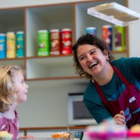 Eine Frau zeigt einem Kind eine Schokoladenform. Beide l&auml;cheln. Im Hintergrund sind bunte Dosen auf Regalen zu sehen., &copy; Alfred Ritter GmbH &amp; Co. KG | Waldenbuch